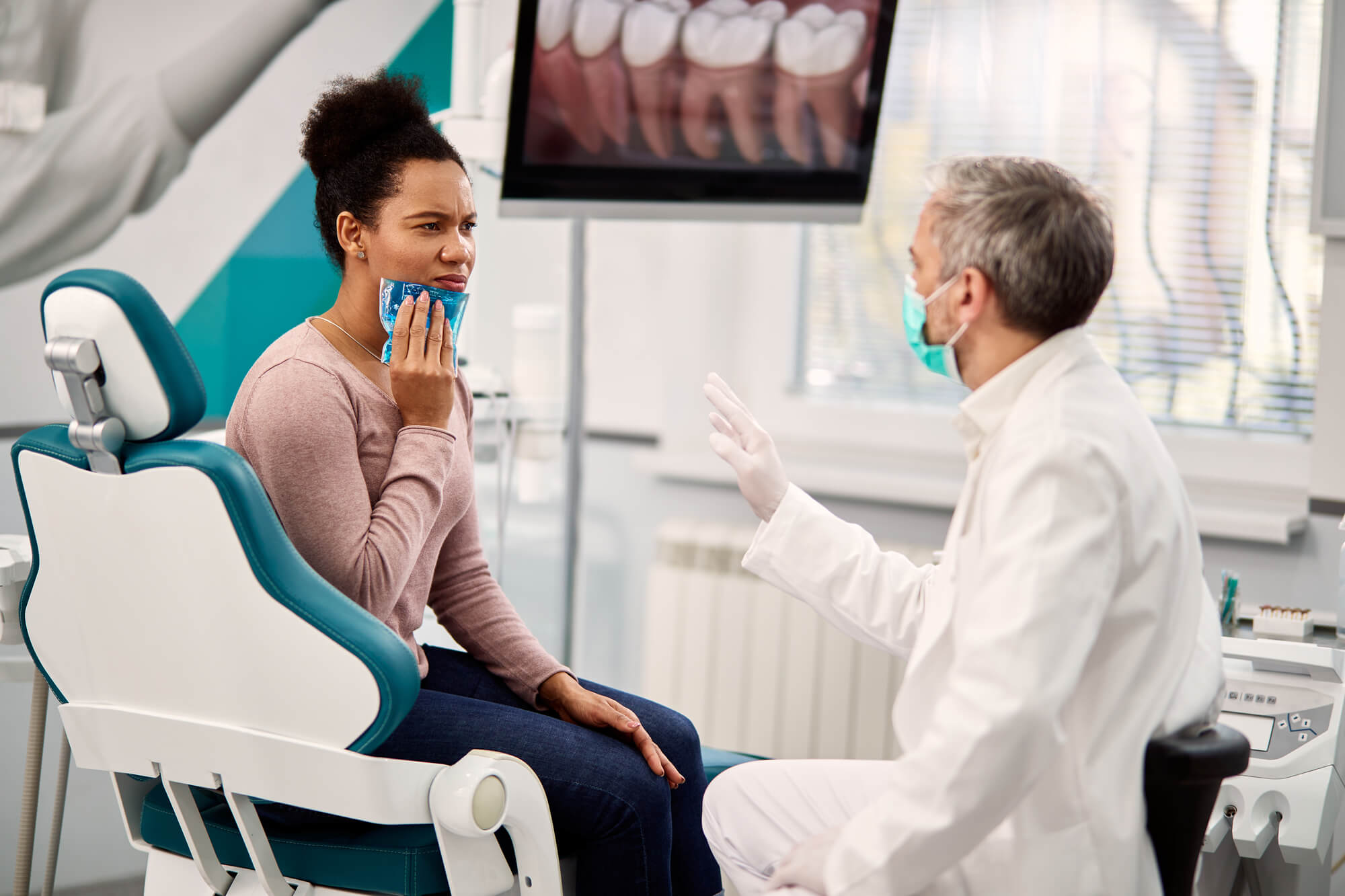 African-American-woman-with-toothache-holding-cooling-gel-on-her-jaw-while-communicating-with-stomatologist-during-dental-check-up-at-dentists-office